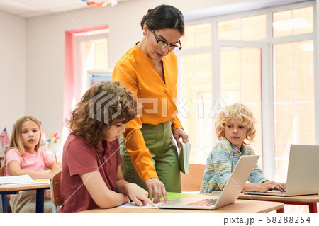 Elegant female teacher explaining work on laptop to children Elegant female teacher explaining work on laptop to children 68288254