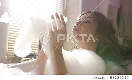 Young attractive woman blowing foam and playing with it. Video. Sun shining through the window in the bathroom where brunette taking bubble bath and relaxing. Young attractive woman blowing foam and playing with it. Video. Sun shining through the window in the bathroom where brunette taking bubble bath and relaxing. 68289219