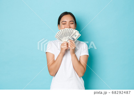 Shopping, money and finance concept. Young successful and lucky cheerful asian girl smelling dollars and smiling satisfied, enjoying being rich, holding cash over light-blue background Shopping, money and finance concept. Young successful and lucky cheerful asian girl smelling dollars and smiling satisfied, enjoying being rich, holding cash over light-blue background 68292487