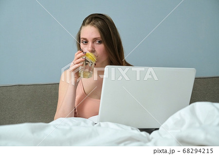 Young woman sitting in bed with laptop computer and drinking water with lemon Young woman sitting in bed with laptop computer and drinking water with lemon 68294215