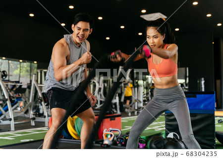 Asian personal trainer coaching a bodybuilding woman to perform crossfit battle ropes exercise in the fitness gym Asian personal trainer coaching a bodybuilding woman to perform crossfit battle ropes exercise in the fitness gym 68304235
