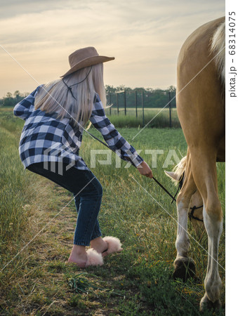 young blonde girl dressed in a hat and funny shoes pulls a horse on a farm in the village 68314075