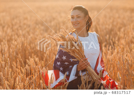 Woman with American flag and with a sheaf of ears in wheat field at sunset. 4th of July. Independence Day patriotic holiday. 68317517