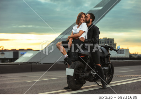 Young sexy woman hugging cute man in stylish black leather jacket, sitting on sports motorcycle on the bridge in the city on sunset and kissing 68331689