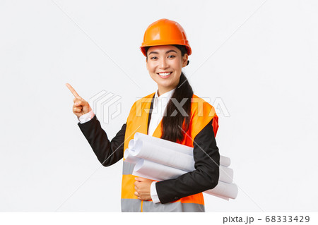 Smiling professional asian female architect, engineer in safety helmet introduce construction project, pointing hand left as carry blueprint, giving speech, introduce chart or plan of building 68333429