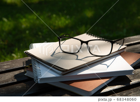glasses lie on top of a pile of books with a notebook on a bench in the park  68348060