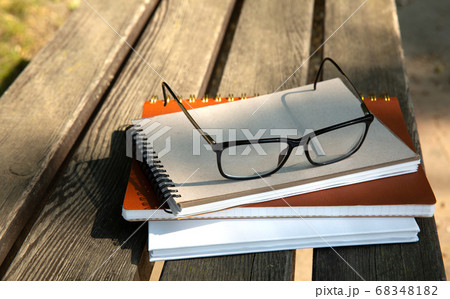 glasses lie on top of a pile of books with a notebook on a bench in the park  68348182