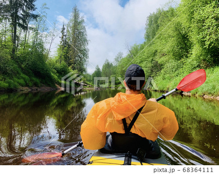 The men in a cap and raincoat of orange color floats on a kayak on the forest quiet river, the beautiful landscape, a changeable weather, actively rows with an oar, beautiful reflection The men in a cap and raincoat of orange color floats on a kayak on the forest quiet river, the beautiful landscape, a changeable weather, actively rows with an oar, beautiful reflection 68364111