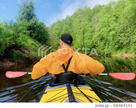 The men in a cap and raincoat of orange color floats on a kayak on the forest quiet river, the beautiful landscape, a changeable weather, actively rows with an oar, beautiful reflection The men in a cap and raincoat of orange color floats on a kayak on the forest quiet river, the beautiful landscape, a changeable weather, actively rows with an oar, beautiful reflection 68364141