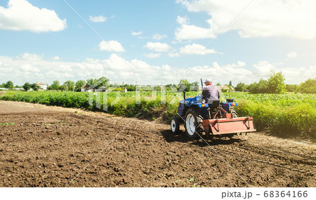 Farmer drives a tractor with a milling machine. Loosening surface, cultivating the land. Farming, agriculture. Loosens, grind and mix soil on plantation field. Field preparation for new crop planting. Farmer drives a tractor with a milling machine. Loosening surface, cultivating the land. Farming, agriculture. Loosens, grind and mix soil on plantation field. Field preparation for new crop planting. 68364166