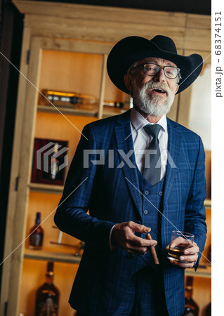 Portrait of old-aged male dandy in big hat smokes cigar and drin 68374151