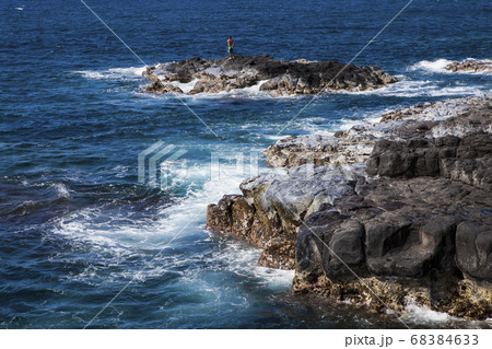 Black laval rocks on a jagged coastline with waves breaking, one person standing on an outcrop. 68384633