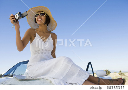 Native American woman in sun dress sitting on white convertible sports car, taking pictures Native American woman in sun dress sitting on white convertible sports car, taking pictures 68385355