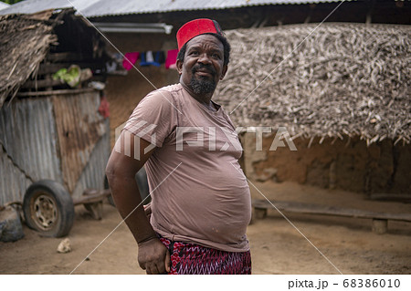 An African Older Man in Red Muslim Taqiyyah Fez Hat posing with a stick for lame people on Yard Near the Basic Hut with Thatched roof in Small Remote Village in Tanzania, Pemba island, Zanzibar 68386010