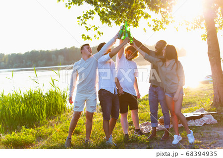 Group of friends clinking beer bottles during picnic at the beach. Lifestyle, friendship, having fun, weekend and resting concept. Group of friends clinking beer bottles during picnic at the beach. Lifestyle, friendship, having fun, weekend and resting concept. 68394935