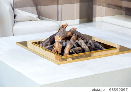 open fireplace in the center of the living room with a stone worktop and a glass chimney body, close up of the fireplace with firewood and a gold frame. 68399067