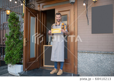 Bearded waiter in apron standing at the door, holding open sign 68410213