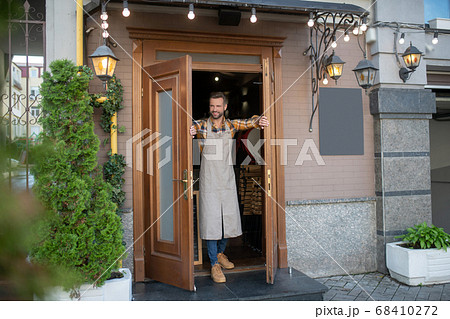 Young bearded male in grey apron opening cafe doors 68410272