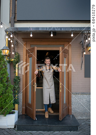 Bearded male in apron opening cafe doors, smiling Bearded male in apron opening cafe doors, smiling 68410279