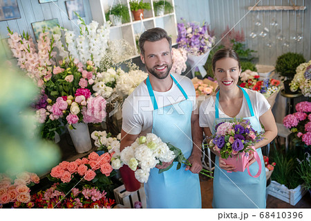 Bearded male and brown-haired female in aprons, holding bouquet and box of flowers, smiling Bearded male and brown-haired female in aprons, holding bouquet and box of flowers, smiling 68410396