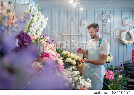 Smiling bearded young male in apron spraying roses in front of flower stand Smiling bearded young male in apron spraying roses in front of flower stand 68410454