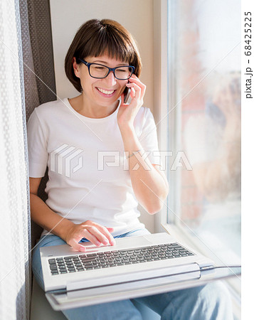 Woman with smartphone remote works from home. She smiles widely  on window sill with laptop on knees. Lockdown quarantine because of coronavirus COVID19. Self isolation at home. 68425225