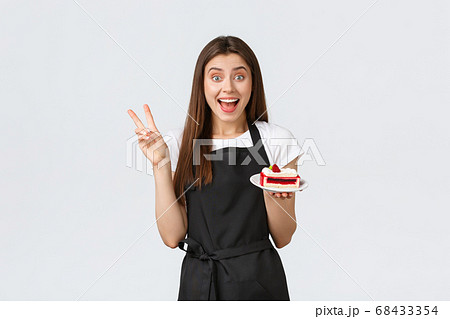 Grocery store employees, small business and coffee shops concept. Excited cheerful female cafe worker in black apron smiling amused and showing peace gesture as serving delicious cake in cafe Grocery store employees, small business and coffee shops concept. Excited cheerful female cafe worker in black apron smiling amused and showing peace gesture as serving delicious cake in cafe 68433354