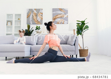 Happy calm mother doing morning exercises in yoga pose while her little daughter playing at home. 68434781