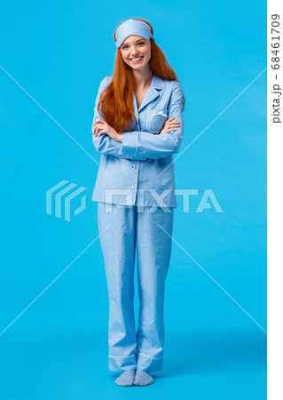 Vertical full-length shot happy cheerful caucasian foxy woman, teenage redhead girl in nightwear and sleep mask, standing blue background, cross arms over chest smiling 68461709