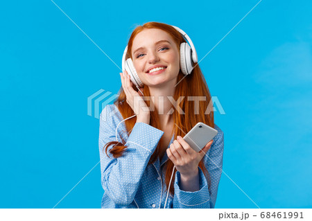 Technology, music and entertainment concept. Cheerful attractive redhead caucasian woman listening music in headphones, holding smartphone, smiling delighted, standing blue background Technology, music and entertainment concept. Cheerful attractive redhead caucasian woman listening music in headphones, holding smartphone, smiling delighted, standing blue background 68461991
