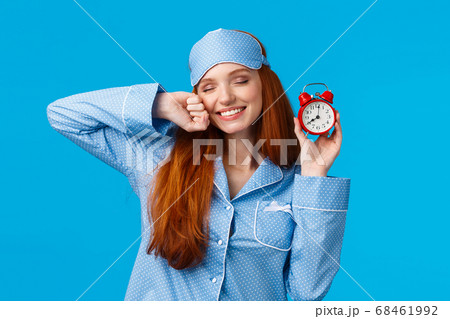 Girl slept well, wake up energized, sleepy stretching with closed eyes and lovely smile, holding red cute clock, set up alarm to be work in time, wearing pyjama and sleep mask, blue background Girl slept well, wake up energized, sleepy stretching with closed eyes and lovely smile, holding red cute clock, set up alarm to be work in time, wearing pyjama and sleep mask, blue background 68461992