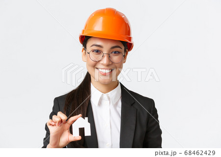 Close-up of happy asian female engingeer, real estate agent in helmet and business suit holding house miniature and smiling, architect working over renovation project, white background Close-up of happy asian female engingeer, real estate agent in helmet and business suit holding house miniature and smiling, architect working over renovation project, white background 68462429