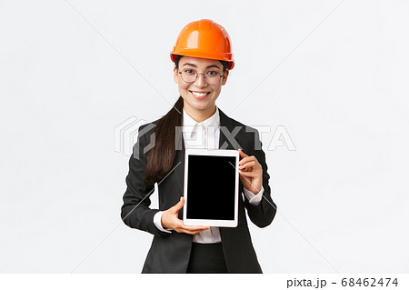 Smiling professional female engineer, saleswoman or estate agent showing location to client, standing in safety helmet and business suit, introduce construction project at tablet screen Smiling professional female engineer, saleswoman or estate agent showing location to client, standing in safety helmet and business suit, introduce construction project at tablet screen 68462474