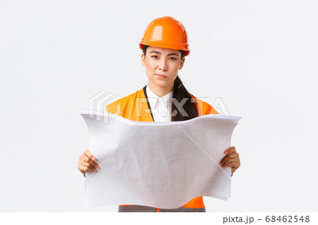 Thoughtful professional asian female construction manager, architect reading blueprints and looking at camera pondering, making decision while studying project plan, white background 68462548