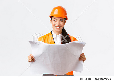 Creative asian female architect imaging her plan, standing in safety helmet and looking away thoughtful while reading blueprints, picturing buiding after finish construction works, white background 68462953