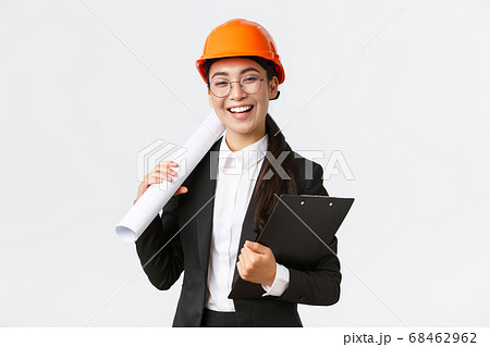 Professional happy asian female architect, construction engineer in helmet and business suit holding blueprints and clipboard with building documentation, smiling cheerful, white background 68462962