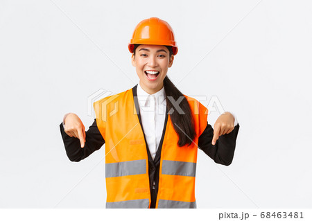 Excited smiling asian female engineer, industrial woman in safety helmet and reflective jacket, pointing fingers down, making announcement. Architect showing her project, white background Excited smiling asian female engineer, industrial woman in safety helmet and reflective jacket, pointing fingers down, making announcement. Architect showing her project, white background 68463481