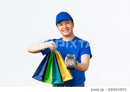 Contactless payment, shopping and home delivery concept. Smiling pleasant asian courier in blue uniform bring client order to doorstep, handing packages and POS terminal for paying 68463955