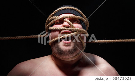 Portrait of binded screaming fat man in cap with rope on face 68473022