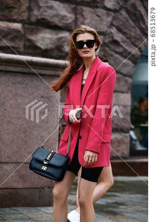 Outdoor close up portrait of young beautiful fashionable girl posing in street. Model wearing stylish sunglasses and bright crimson oversized blazer with black leather bag. City lifestyle. Female 68513679