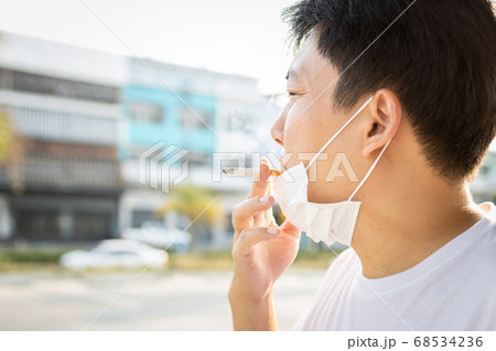 Asian man with a protective mask during Covid-19 pandemic,hold a cigarette in his mouth,smokers male smoking,spreading of Coronavirus with droplets come out from smoke of cigarette,airborne diseases 68534236