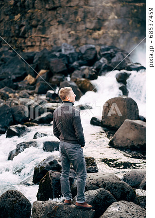 A guy in a black knitted sweater stands on rocks near Ehsaraurfoss Falls, Ehsarau River, National Park, Tingvedlir, Syudurland Region, Iceland. A guy in a black knitted sweater stands on rocks near Ehsaraurfoss Falls, Ehsarau River, National Park, Tingvedlir, Syudurland Region, Iceland. 68537459
