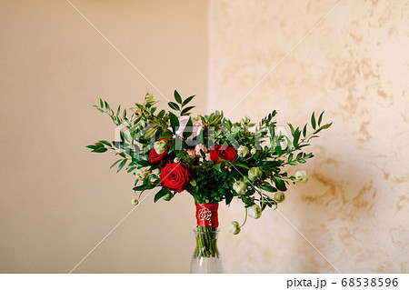 bridal bouquet of red and pink roses, boxwood branches, not blooming buds of white flowers and red ribbons with brooch in the glass vase 68538596