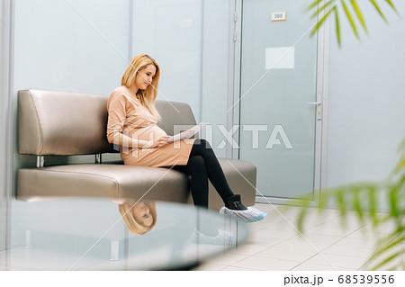 Smiling young pregnant woman is sitting in hallway of hospital waiting to see gynecologist. Smiling young pregnant woman is sitting in hallway of hospital waiting to see gynecologist. 68539556