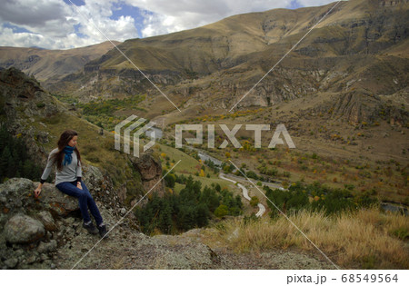 Young woman with long hair sitting on stone on background of mountains and brown valley with river, view from side 68549564