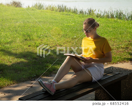 A beautiful young woman student in glasses, reading a book, sits on a bench 68552416