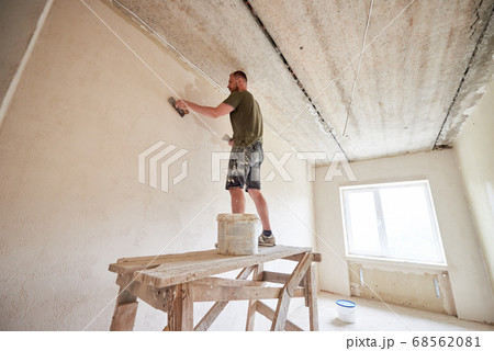 Young man is standing on wooden stand and working with spatula with plaster at the wall against a window. 68562081