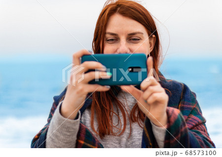 Tourist caucasian red hair woman smiles, takes pictures or recording video on the phone on the beach near the ocean, vacation concept 68573100