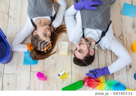 Tired man and woman lie on floor and look at each other in living room with cleaning supplies 68580533