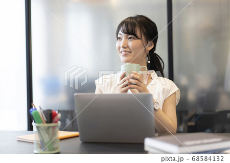オフィスの席でパソコンの前に座り両手でマグカップを持つ女性の写真素材